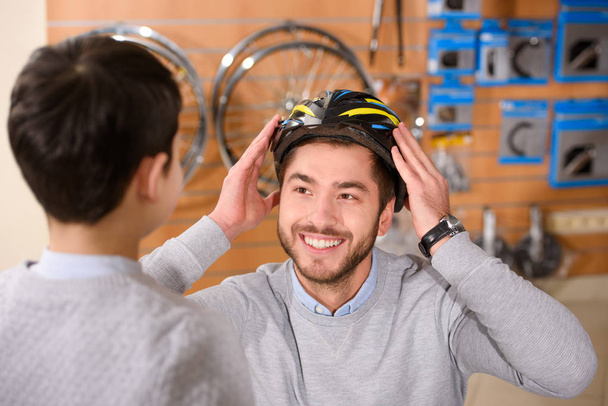 boy looking at happy father wearing bicycle helmet in bike shop - Foto, Imagen