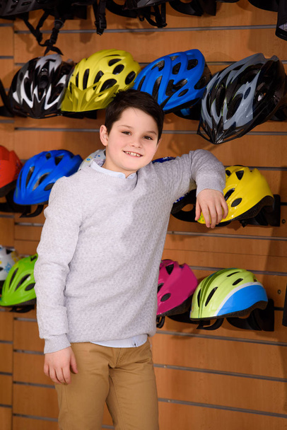 cute little boy leaning at bicycle helmets and smiling at camera in bike shop - Photo, Image