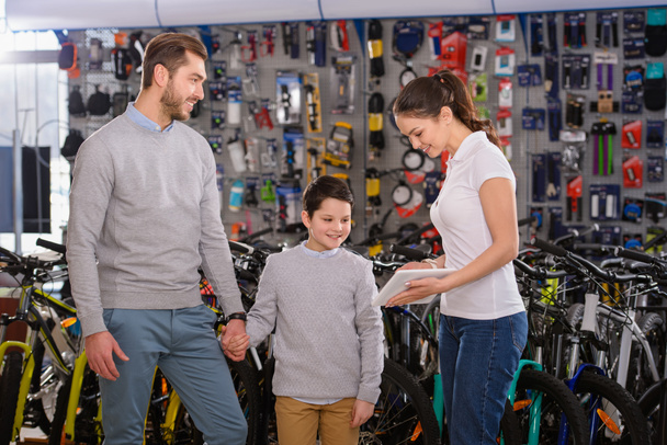 joven sonriente mostrando tableta digital al padre y al hijo eligiendo bicicletas en la tienda de bicicletas
 - Foto, Imagen