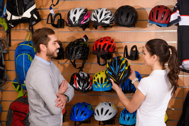 young female manager showing bicycle helmet to smiling customer in bike shop - Fotoğraf, Görsel