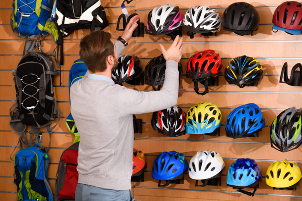 вид сзади молодого человека, выбирающего велосипедные шлемы в магазине велосипедов
 - Фото, изображение