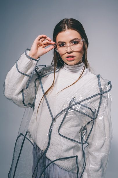 portrait of young woman in fashionable raincoat and eyeglasses looking at camera on grey background - Foto, afbeelding