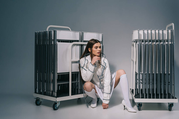 stylish woman in white clothing and raincoat with collapsible chairs behind looking away on grey background  - Фото, зображення