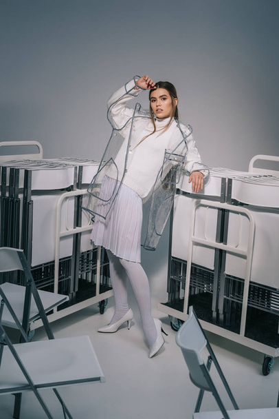 stylish woman in white clothing and raincoat posing with collapsible chairs behind on grey background  - Fotografie, Obrázek