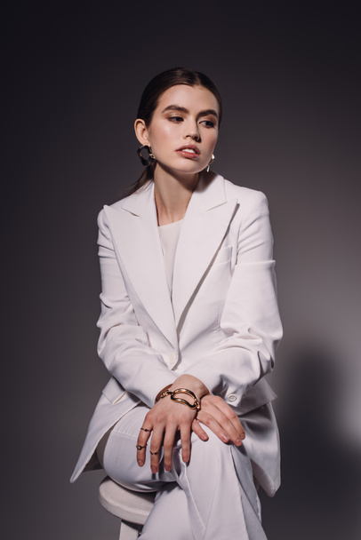 portrait of fashionable woman in white suit sitting on chair on dark background - Fotó, kép