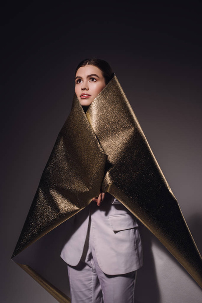 portrait of fashionable pensive woman in golden wrapping paper looking away on dark backdrop - Foto, Imagem
