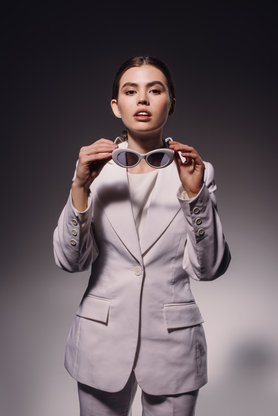 portrait of stylish woman in white suit and eyeglasses posing on dark backdrop - Foto, afbeelding