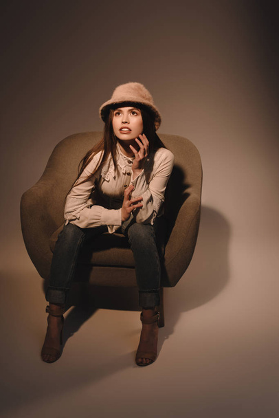 fashionable pensive woman in hat sitting in armchair on dark backdrop - 写真・画像