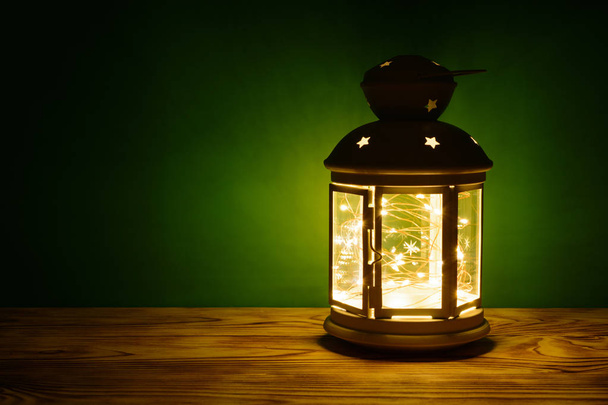 A lantern with a glowing garland in on the table, green background - Photo, Image