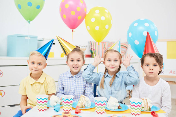Wiersz z śmieszne dzieci w urodziny czapki siedzi przez świąteczny stół kawałki, jeśli ciasto, napoje i ciasteczka - Zdjęcie, obraz