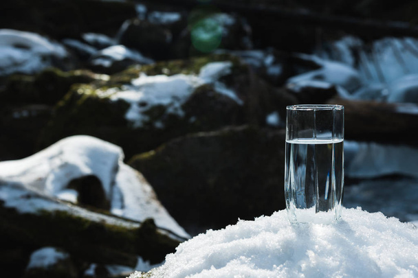A transparent glass glass with drinking mountain water stands in the snow against a background of a clean frost mountain river in winter. - Fotografie, Obrázek