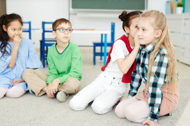 Groep van jonge klasgenoten fluisteren elkaar nieuwsgierig zin of zinsnede terwijl het spel van de vrije tijd - Foto, afbeelding