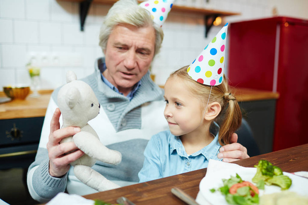 Granddaughter in birthday cap looking at teddybear held by her grandfather while playing by table - Photo, Image