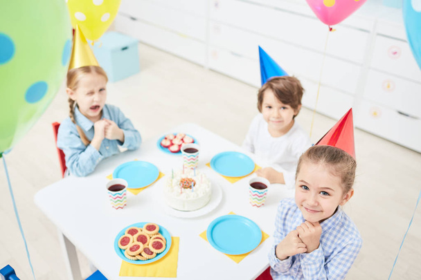 Симпатичні маленькі дівчата і хлопчик сидять за столом на день народження в дитячому саду і розважаються
 - Фото, зображення