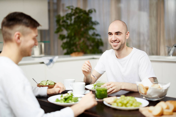 Happy young homosexual couple sitting by table in the kitchen and having breakfast - Foto, afbeelding