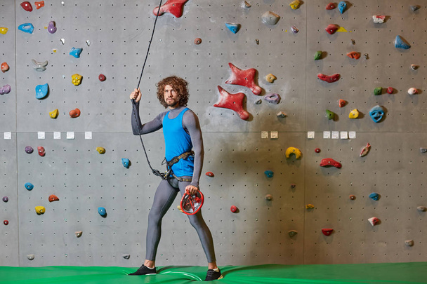 Young sporty man holding by rope during training against climbing wall - Photo, Image