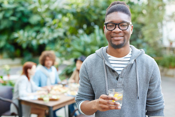 ragazzo africano con rinfrescante bere guardando fotocamera con gli amici a cena su sfondo
 - Foto, immagini