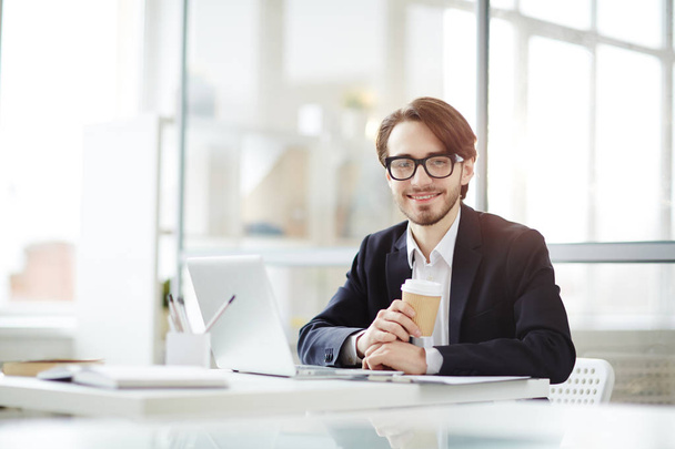 Успешный бизнесмен с напитком, смотрящий в камеру во время кофе-брейка, сидя на рабочем месте перед ноутбуком
 - Фото, изображение
