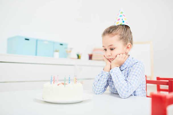 Грустная маленькая девочка смотрит на праздничный торт со свечами, сидя одна за столом
 - Фото, изображение