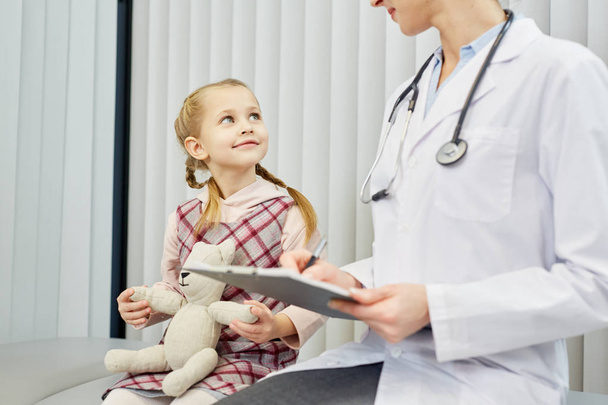 Menina feliz com ursinho escutando o conselho de seu médico em clínicas
 - Foto, Imagem