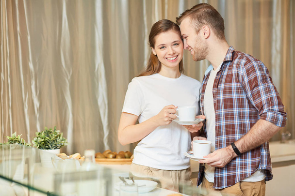 junger, zärtlicher Mann, der neben seiner Frau steht, während er in der Küche Morgentee trinkt - Foto, Bild