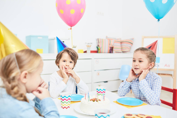 Lindos amiguitos en gorras de cumpleaños reunidos por la mesa festiva con sabrosa torta en su centro
 - Foto, imagen