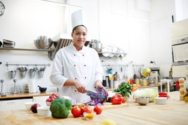 junge Frau in Kochuniform kocht frischen Gemüsesalat, während sie lila Kohl schneidet - Foto, Bild