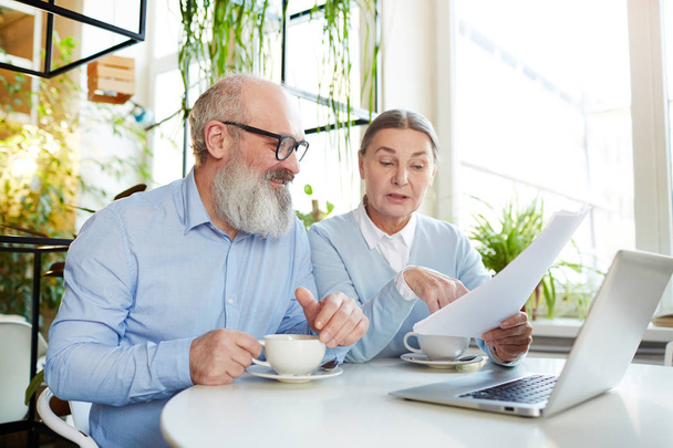 Kaksi kypsää liikemiestä älykkäässä rento istuu pöydän ääressä kahvilassa kannettavan tietokoneen edessä ja keskustelee paperista
 - Valokuva, kuva