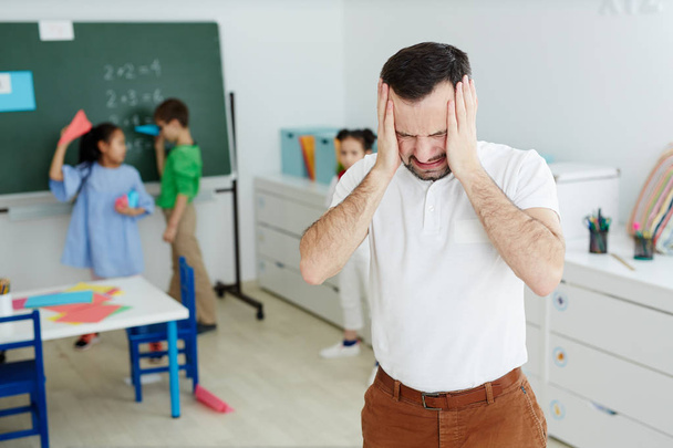 Profesor molesto tocándose la cabeza durante el descanso entre clases mientras grupo de niños se divierten en el fondo
 - Foto, Imagen