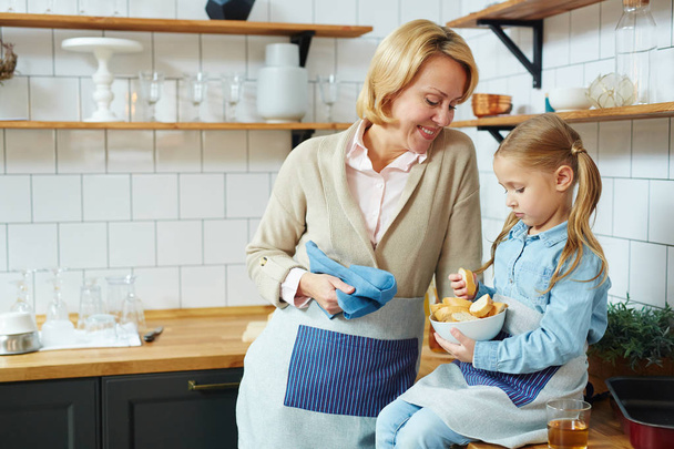 Onnellinen kypsä nainen ja hänen pieni tyttärentytär kulhoon vehnäleipää istuu keittiössä
 - Valokuva, kuva