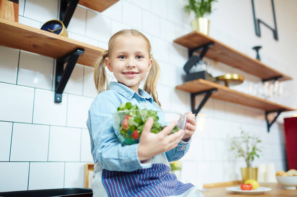 Smiling little girl with bowl of fresh vegetable salad looking at camera in the kitchen - 写真・画像