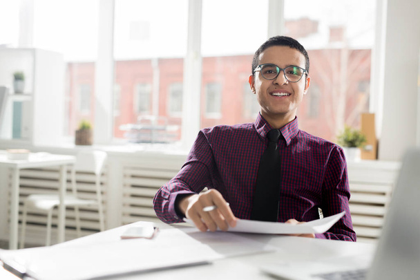 glücklicher junger Geschäftsmann in Brille, Hemd und Krawatte, der am Schreibtisch im Büro mit Papieren arbeitet - Foto, Bild