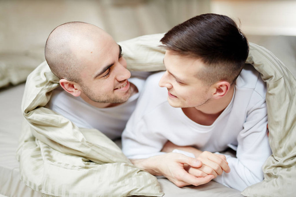 Two amorous guys lying on bed under blanket and lookign at one another with smiles - Fotó, kép