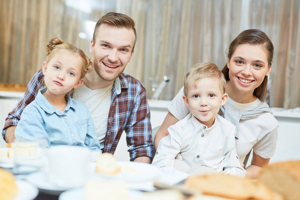 Funny siblings and their young parents looking at camera while sitting by table - Photo, Image