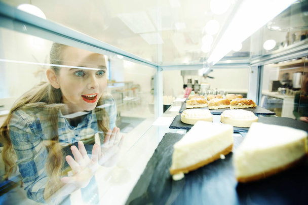 Amazed girl looking at tasty pastry through display glass in cafeteria - Photo, Image