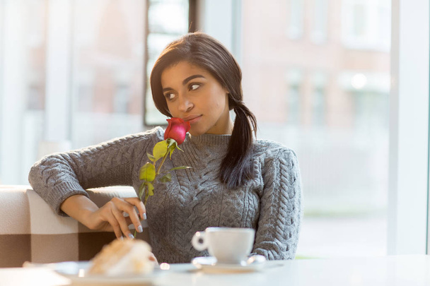 Fiatal nő a szürke pulóver, illatú friss piros rózsa, miközben időt kávézó - Fotó, kép
