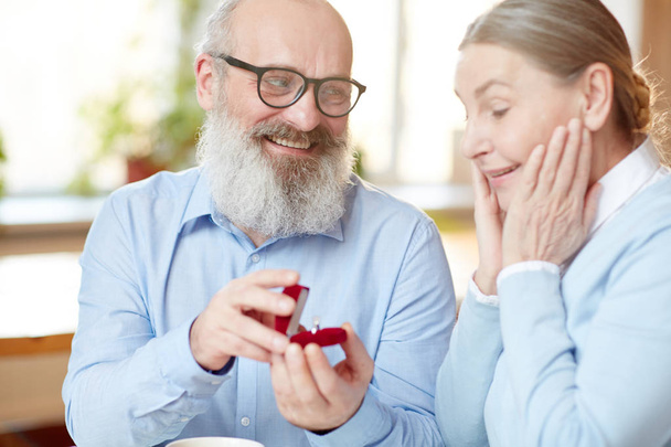 glücklicher älterer Mann blickt überraschte Frau an und zeigt ihren Diamantring in Samtbox - Foto, Bild