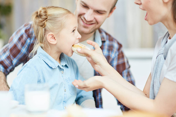 Jeune mère prudente nourrissant sa fille avec un beignet frais et savoureux au petit déjeuner avec son père proche
 - Photo, image