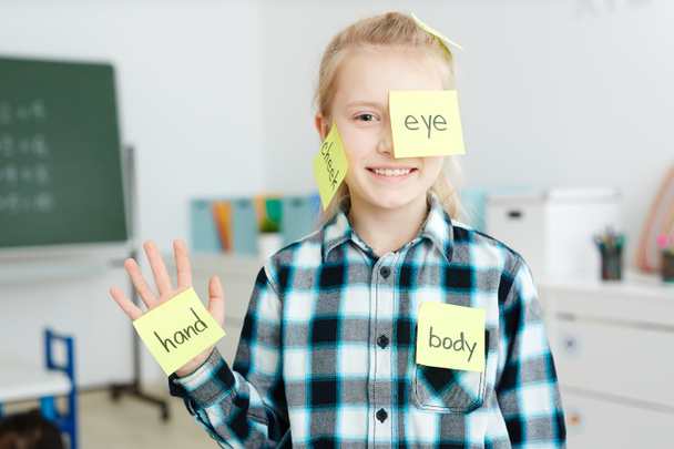 Happy schoolgirl having sticky note papers with English names of body parts on her hand, cheek and eye - Photo, Image