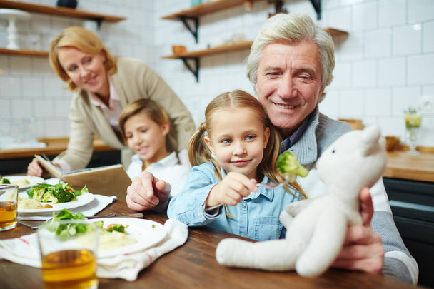 Happy mature man holding teddybear while his granddaughter giving food to her toy by dinner - Photo, Image