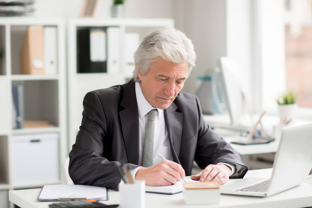 Senior businessman making notes in notebook while sitting by workplace in office - Photo, Image