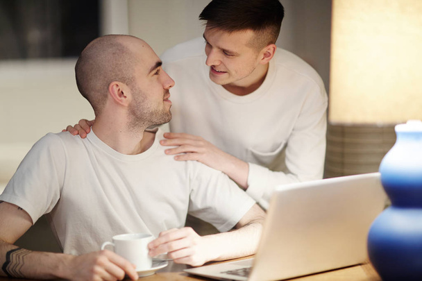 Amoureuze jonge homoseksuele man met kopje drinken en laptop kijken naar zijn geliefde omarmen hem - Foto, afbeelding