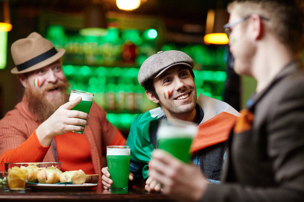 Homme souriant avec un verre de bière en écoutant l'un des compagnons pendant la conversation tout en étant assis dans le bar
 - Photo, image
