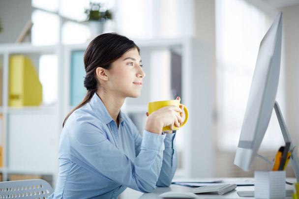 Mujer de negocios contemporánea con taza de café pasar tiempo delante del monitor de computadora por el lugar de trabajo
 - Foto, Imagen