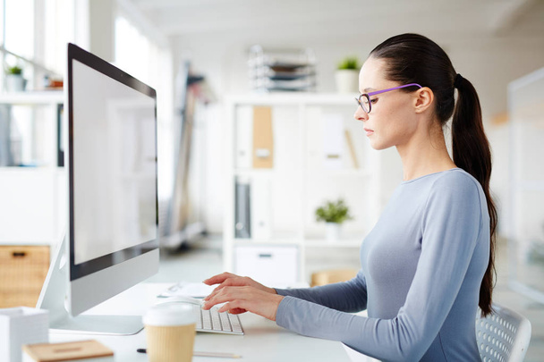 Молодая серьезная женщина в повседневной одежде сидит за столом перед монитором компьютера и вводит данные
 - Фото, изображение