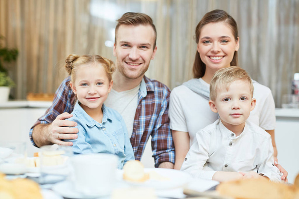 Felices padres jóvenes abrazando a su lindo hijo y su hija mientras están sentados junto a la mesa servida
 - Foto, Imagen