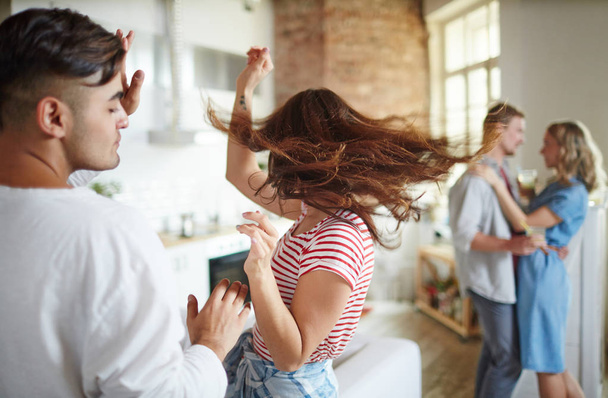 Chica extática sacudiendo su pelo largo mientras baila en la fiesta en casa con su novio cerca y pareja amorosa en el fondo
 - Foto, imagen
