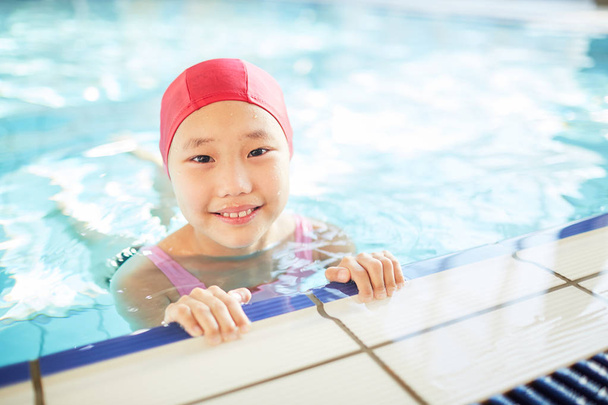 Nettes kleines Mädchen mit Badekappe blickt beim Training im Schwimmbad in die Kamera - Foto, Bild