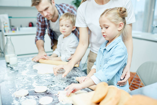 Современная семья из четырех человек смешивает и скатывает тесто вместе во время приготовления домашней выпечки на кухне
 - Фото, изображение