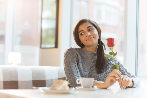 Happy young woman with fresh rose sitting by table in cafe and looking aside while thinking about her sweetheart - Foto, Imagem